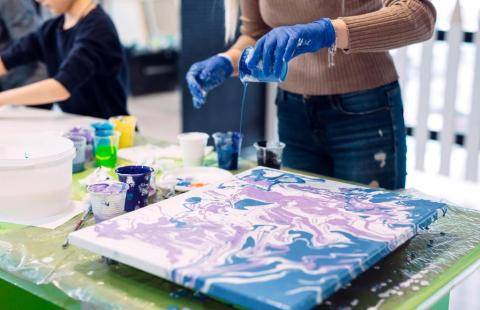 Henkilö kaataa sinistä akryylimaalia kankaalle acrylic pouring -tekniikalla. Pöydällä on useita maalikuppeja, ja valmiissa työssä näkyy sinisen ja vaalean violetin sävyisiä marmorimaisia kuvioita.